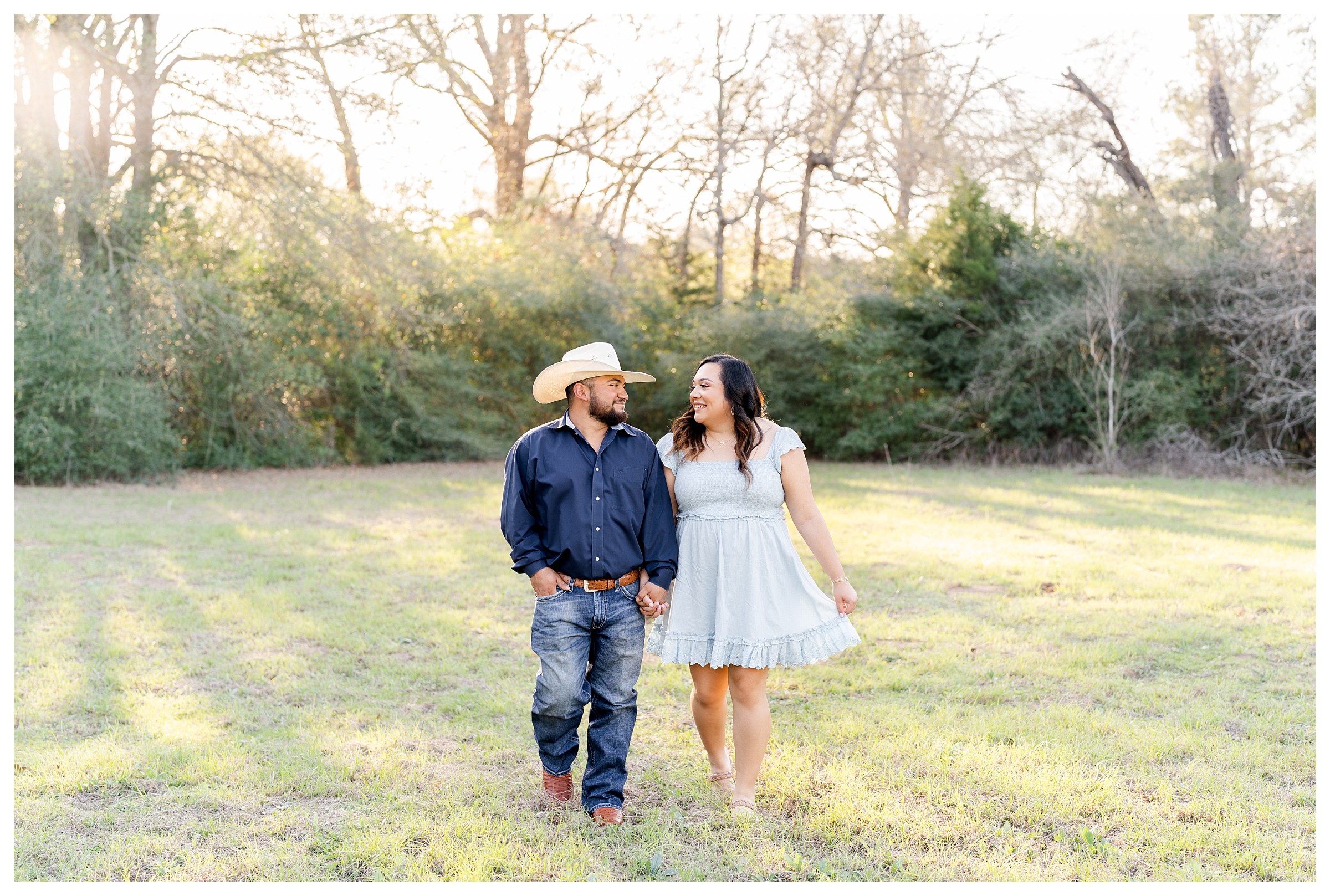 Couple smiling and walking towards camera in an open field at sunset while surrounded by trees and he is wearing a dark blue button down with jeans and cowboy hat while she is wearing a light blue dress