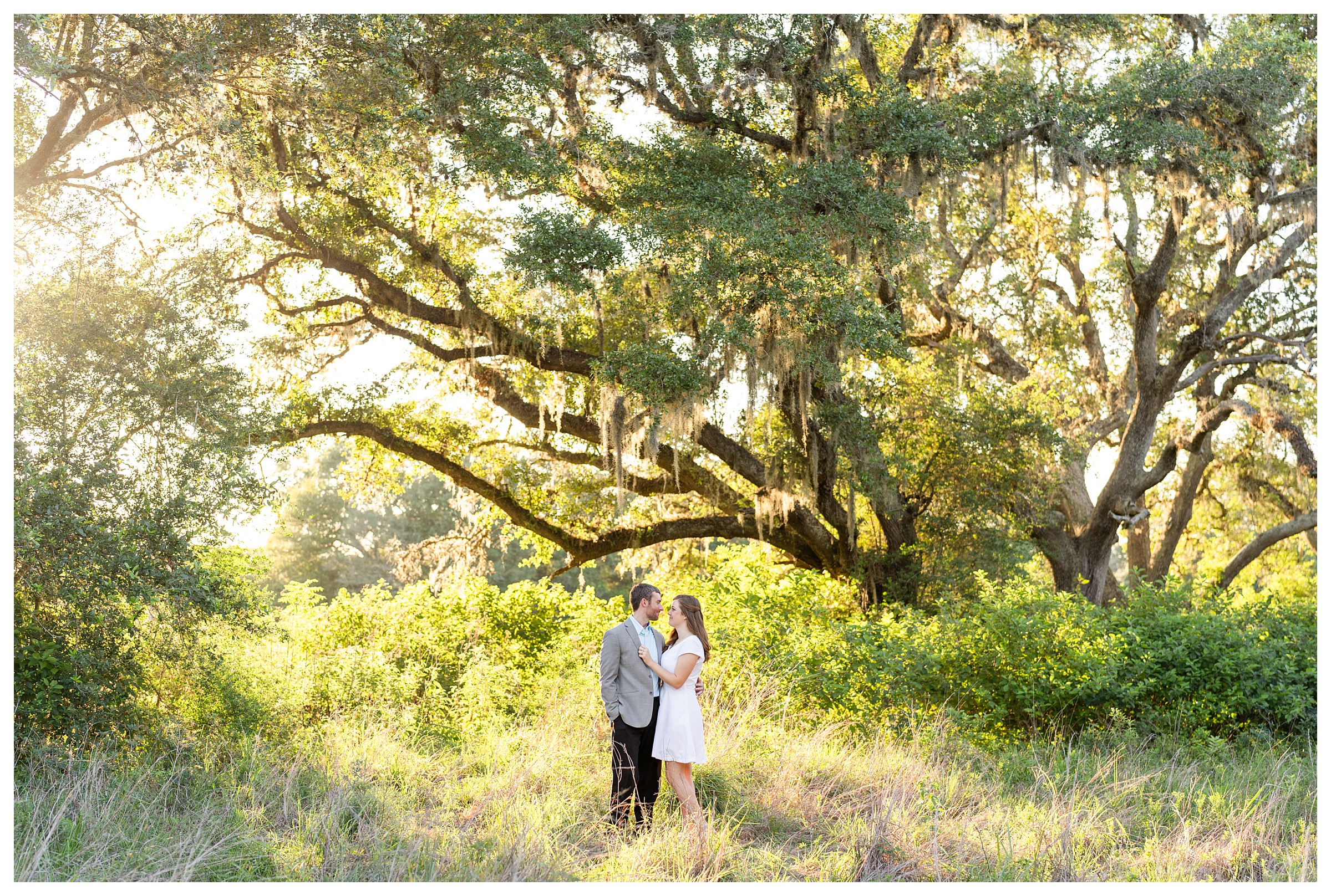 Couple standing belly button to belly button while smiling at each other with her hand on his chest and standing in the middle of a field surrounded by trees and nature with the sun glowing off the trees all around them at Brazos Bend State Park in Houston, Texas