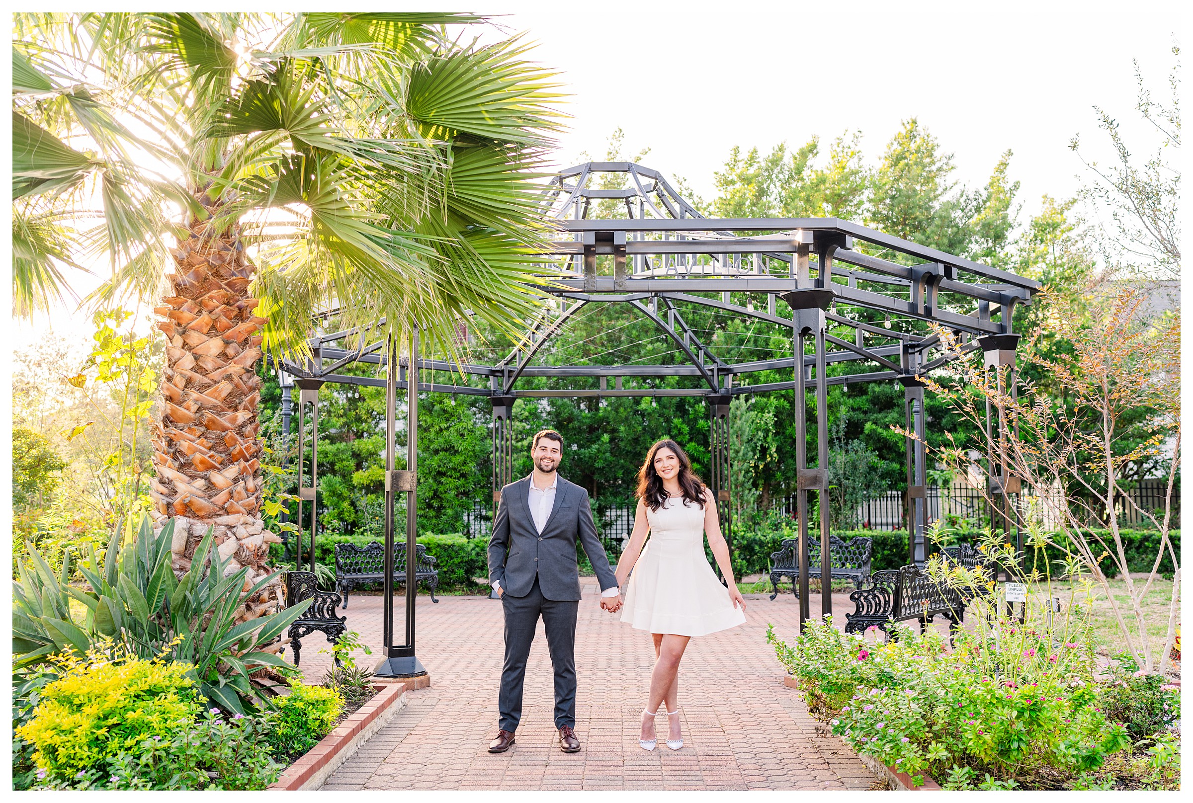 Far away shot of couple standing facing camera while hand in hand and smiling while guy is wearing a dark blue suit and girl is wearing white cocktail length dress with heels in a nature filled park and black iron structures in Galveston, Texas.